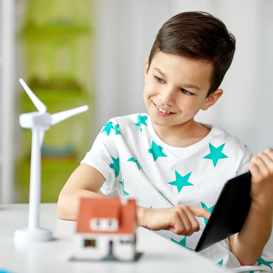 A young boy smiling and holding a tablet while looking at a model wind turbine and a miniature house on a table.